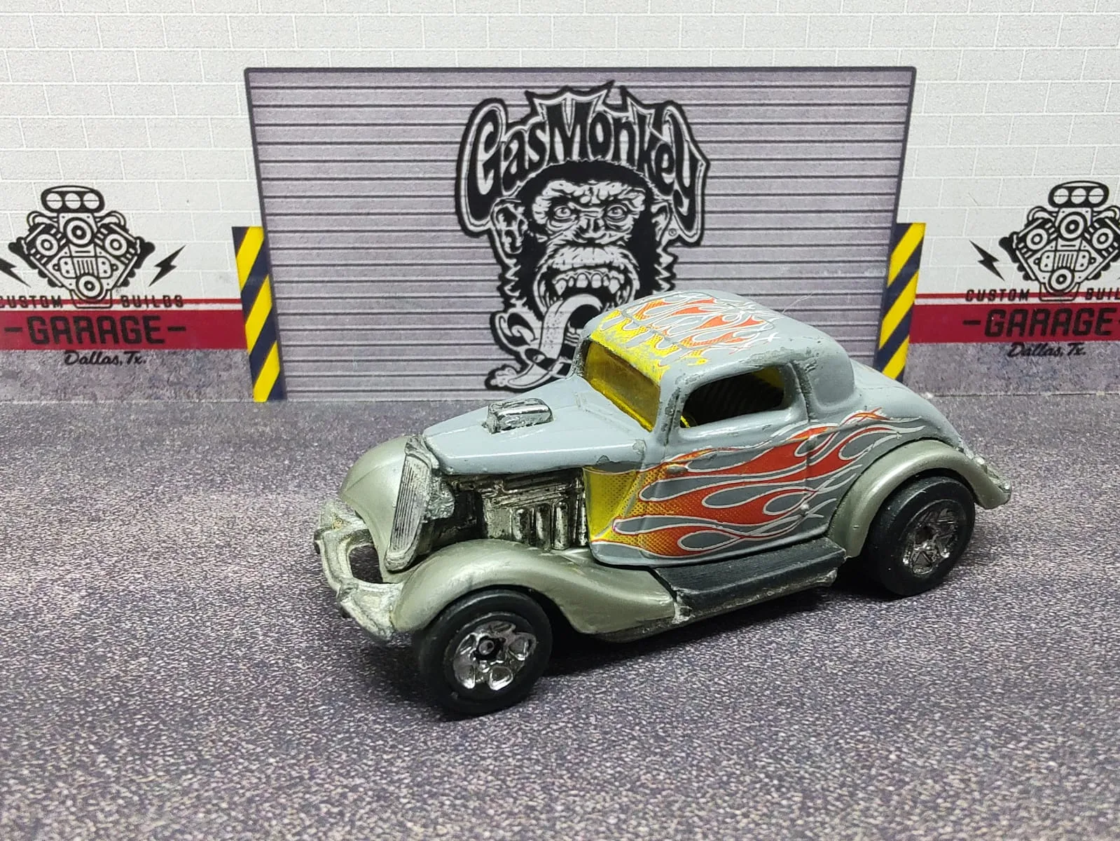 3-Window ’34 — hot rod plateado con llamas rojas y amarillas, motor expuesto y ruedas traseras grandes